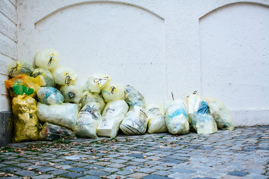 A collection of tightly packed trash bags, primarily made of plastic, with some appearing yellow and others transparent or white, piled against a white textured exterior wall on a cobblestone pavement. The bags vary in size and are filled with waste materials, with some showing visible contents such as packaging or paper. The ground beneath the bags is composed of small, irregularly shaped stones with scattered leaves and debris. The scene is illuminated by natural daylight, casting soft shadows. This setup suggests an outdoor location where waste has been collected for disposal, possibly in connection with private rubbish collection services, such as those provided by Rubbish Collection Belgravia, to avoid hidden rubbish removal charges. The environment is clean and orderly, indicating that the waste is staged for collection or removal, aligning with professional rubbish clearance practices common in urban settings.