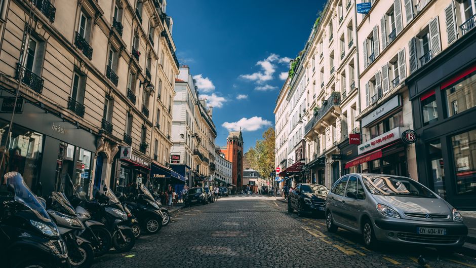 A wide view of a bustling urban street scene showing parked scooters and cars along a cobblestone road lined with multi-story buildings featuring classic European architecture, including ornate window frames and balconies. The street is filled with retail shops and cafes with storefronts displaying signage, some with awnings extending over the sidewalk. Pedestrians are visible walking along the pavement, and a historic red brick church with a pointed steeple is positioned in the background under a clear blue sky with scattered white clouds. The scene appears to be during daytime with natural sunlight illuminating the facades of the buildings, highlighting the varied textures of stone and brick surfaces. The image captures an active city environment where private vehicle and scooter parking is typical, reflecting an area that may involve independent rubbish collection services alongside regular urban waste management. The overall atmosphere is vibrant and lively, characteristic of central city districts with mixed-use commercial and residential properties. Rubbish Collection Belgravia would assess this type of setting for waste removal, particularly in terms of managing waste from shops and offices in such urban environments.