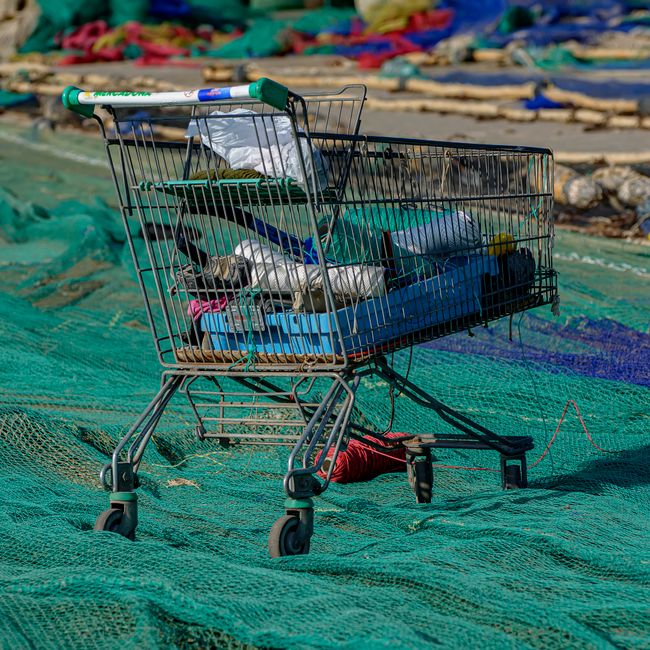 A metal shopping trolley with a plastic handlebar, filled with various discarded items including plastic bottles, paper, and miscellaneous waste, is positioned on a large green fishing net stretched across a shoreline or beach area. In the background, out-of-focus colorful fabrics or tarps are visible, suggesting a fishing or market environment. The trolley's structure shows a silver metallic finish with some wear, and it is situated on a surface that appears to be a combination of natural sand and the textured green netting, which is anchored with red and white strings. The scene is illuminated by natural daylight, highlighting the general scene of waste collection or disposal in an outdoor, coastal, or harbour setting, aligning with services provided by companies like Rubbish Collection Belgravia engaged in private waste handling and on-site clearance of non-recyclable rubbish, particularly in areas where waste management occurs outside traditional municipal systems.