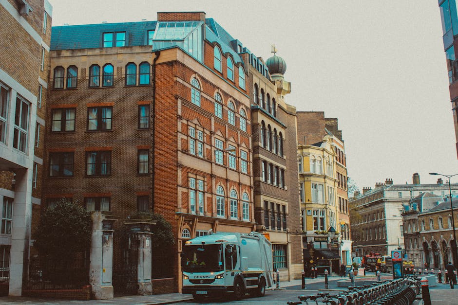 A wide view of a bustling urban street scene showing parked scooters and cars along a cobblestone road lined with multi-story buildings featuring classic European architecture, including ornate window frames and balconies. The street is filled with retail shops and cafes with storefronts displaying signage, some with awnings extending over the sidewalk. Pedestrians are visible walking along the pavement, and a historic red brick church with a pointed steeple is positioned in the background under a clear blue sky with scattered white clouds. The scene appears to be during daytime with natural sunlight illuminating the facades of the buildings, highlighting the varied textures of stone and brick surfaces. The image captures an active city environment where private vehicle and scooter parking is typical, reflecting an area that may involve independent rubbish collection services alongside regular urban waste management. The overall atmosphere is vibrant and lively, characteristic of central city districts with mixed-use commercial and residential properties. Rubbish Collection Belgravia would assess this type of setting for waste removal, particularly in terms of managing waste from shops and offices in such urban environments.