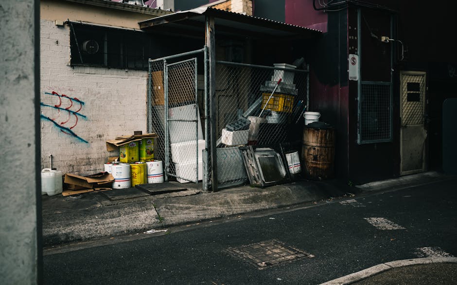 Image depicting an outdoor rubbish storage area located on a paved street, adjacent to a white brick wall with some blue graffiti. The area is enclosed by a metal chain-link fence and contains various discarded items including large white plastic containers, cardboard boxes, metal trays, and a wooden barrel. There are also smaller items such as plastic bins, buckets, and a white bowl stacked or leaning against the fence and nearby structures. The surface is a rough concrete pavement with visible wear, and the background features a dark building with a metal door and a small window. The scene appears to be part of a private waste collection or on-site rubbish disposal setup, handled by Rubbish Collection Belgravia, fitting within the context of alternative waste management or rubbish removal services around Elizabeth Street Belgravia.
