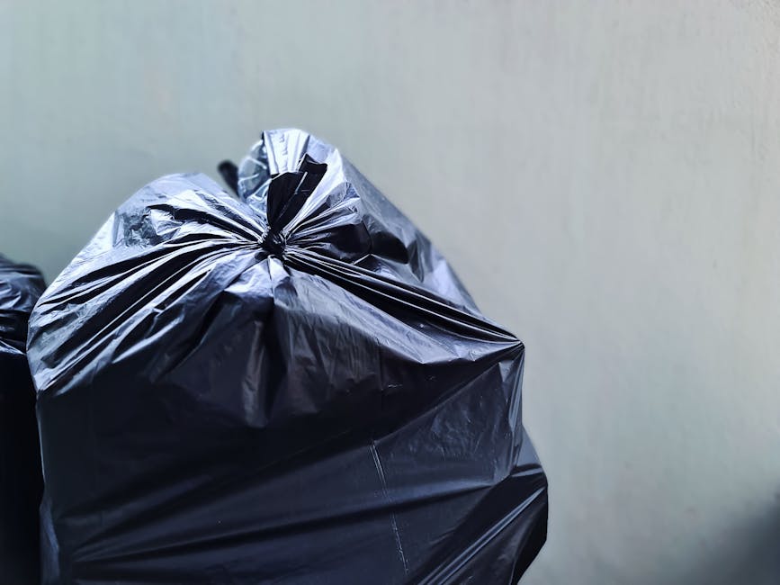 Close-up view of a tightly tied black plastic rubbish bag made of glossy, rippled material, placed in front of a plain, off-white wall. The bag appears to be full, with visible creases and folds indicating its contents. In the background, a partially visible second black rubbish bag is slightly blurred, suggesting an accumulation of waste awaiting collection. The scene is illuminated by natural or overhead lighting, creating gentle reflections on the plastic surface. The setting indicates an indoor or a private outdoor space, such as a driveway or service area, typical of on-site waste disposal managed by a private rubbish collection service like Rubbish Collection Belgravia. This image visually supports the context of alternative waste handling methods, emphasizing the importance of properly sealed rubbish bags for efficient rubbish removal and disposal services in the Eaton Square SW1X area.