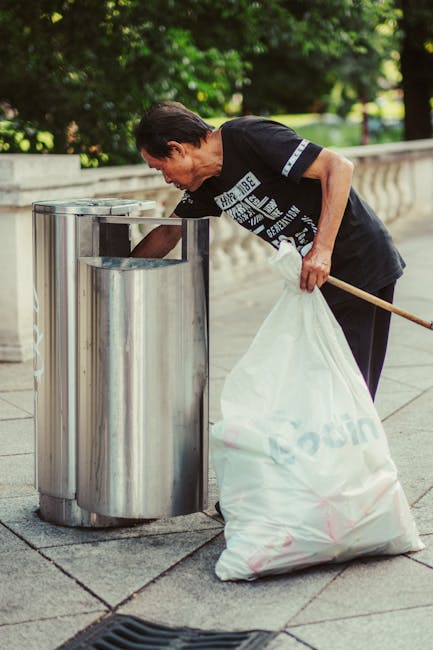 A middle-aged man with dark hair, dressed in a black T-shirt with white text and dark pants, is seen bending over a metallic, stainless steel trash bin on a paved sidewalk. He is holding a large white trash bag with both hands, partially filled with rubbish, and is in the process of emptying or removing it from the bin. The trash bin is cylindrical with a smooth, shiny surface and a partial open top. The scene is set outdoors, with lush green trees and a concrete balustrade visible in the background, indicating a park or urban promenade environment. The lighting appears natural, suggesting daytime, and the overall scene captures a moment of waste disposal or rubbish collection that could relate to private or independent rubbish removal services such as those offered by Rubbish Collection Belgravia, in a setting supporting on-site rubbish handling and waste management.